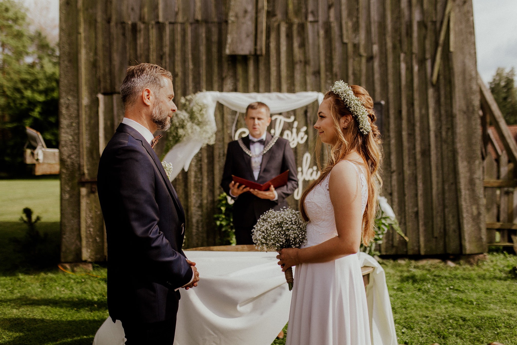 Para młoda stoi przed rustykalnym drewnianym budynkiem podczas ceremonii ślubnej na świeżym powietrzu, z urzędnikiem w tle.