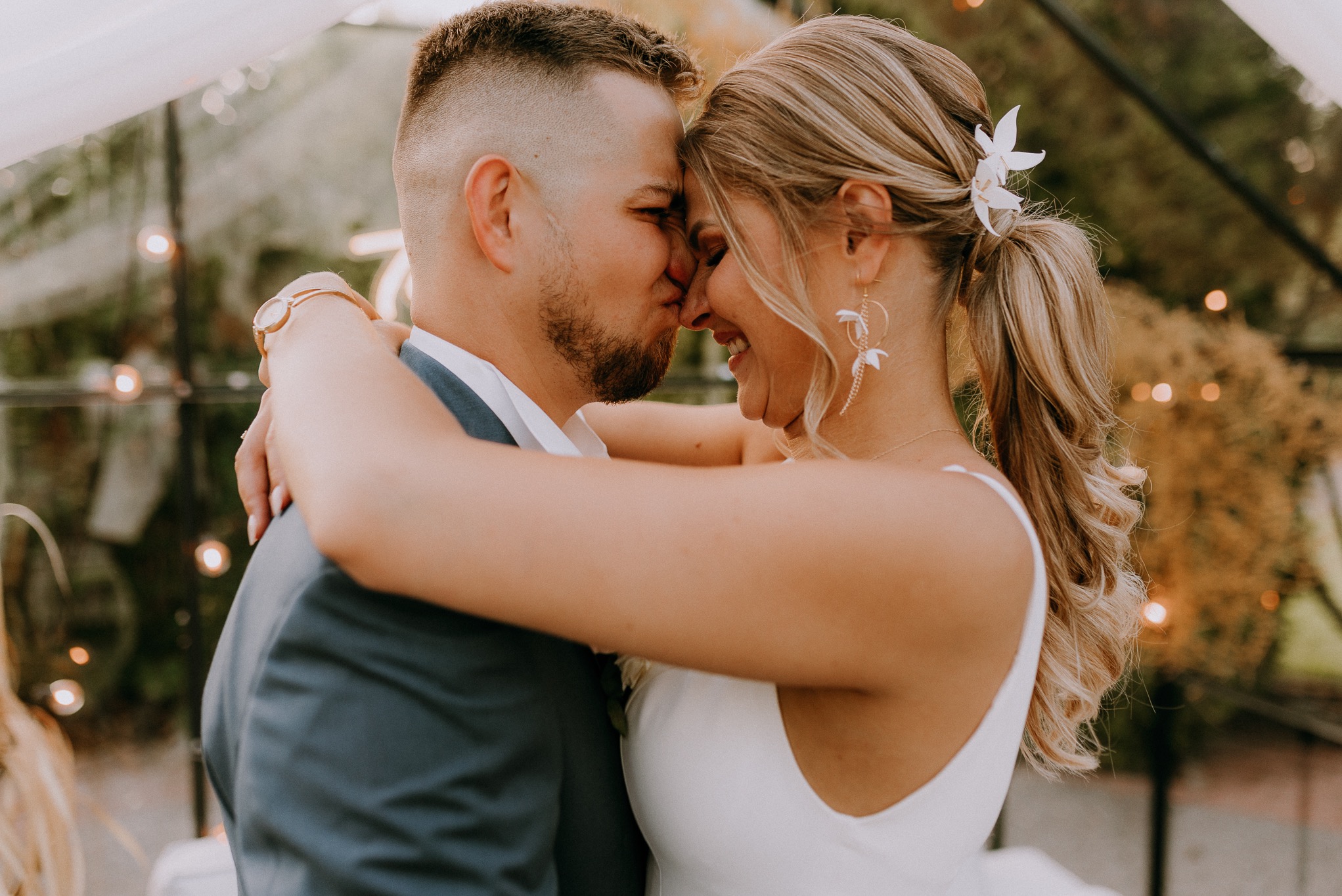 Uśmiechnięta para młoda w objęciach, panna młoda z kwiatem we włosach i delikatnymi kolczykami, pan młody w granatowym garniturze, w tle szklana konstrukcja z zielenią.