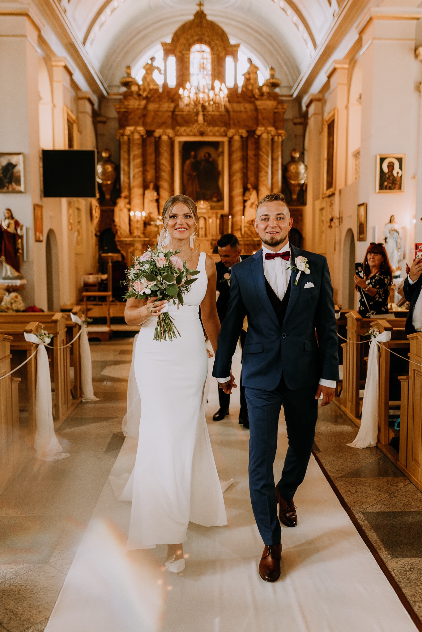 Para młoda, trzymając się za ręce, idzie nawą kościoła po białym dywanie, panna młoda w prostej sukni z bukietem różowych kwiatów, pan młody w granatowym garniturze z bordową muchą.