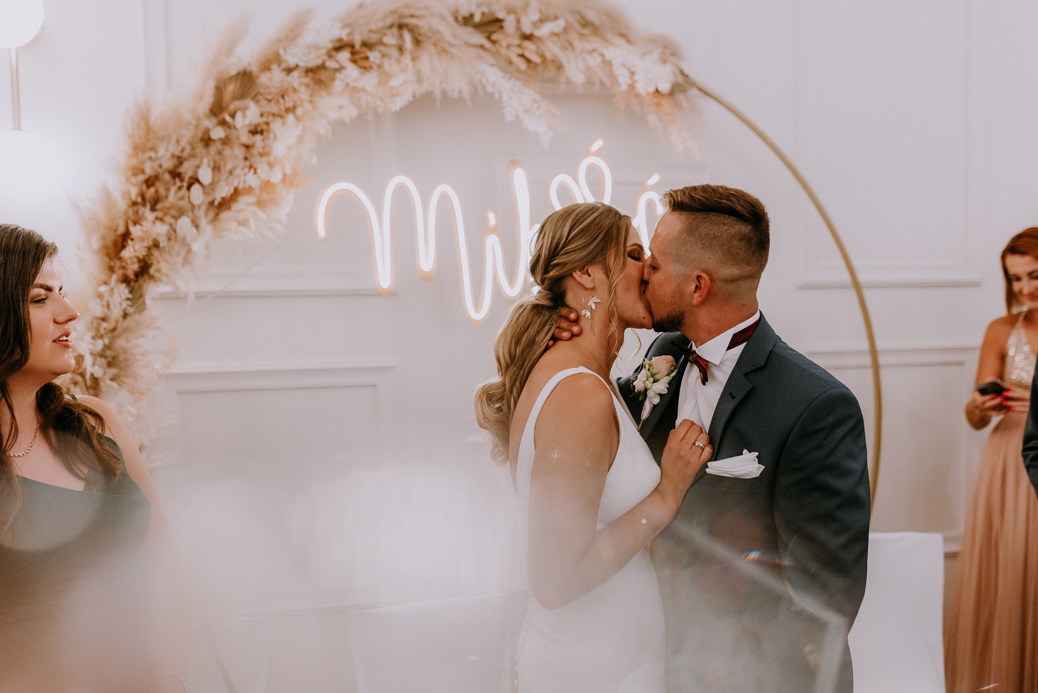 Pocałunek pary młodej na tle dekoracji z pampasów i neonu z napisem. Panna młoda w białej sukni, pan młody w granatowym garniturze z bordową muchą.