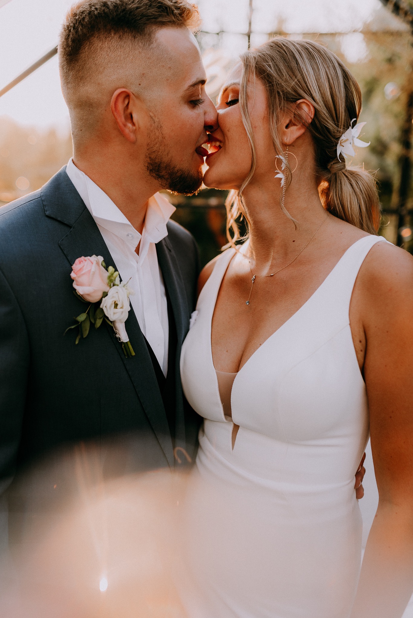 Młoda para w pocałunku, panna młoda w białej sukni z dekoltem w serek i kwiatami we włosach, pan młody w granatowym garniturze z butonierką, ciepłe światło słoneczne.