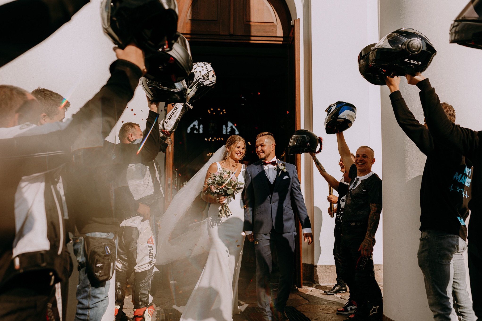 Uśmiechnięta para młoda wychodzi z kościoła, otoczona gośćmi w strojach motocyklowych, unoszącymi kaski w geście świętowania.