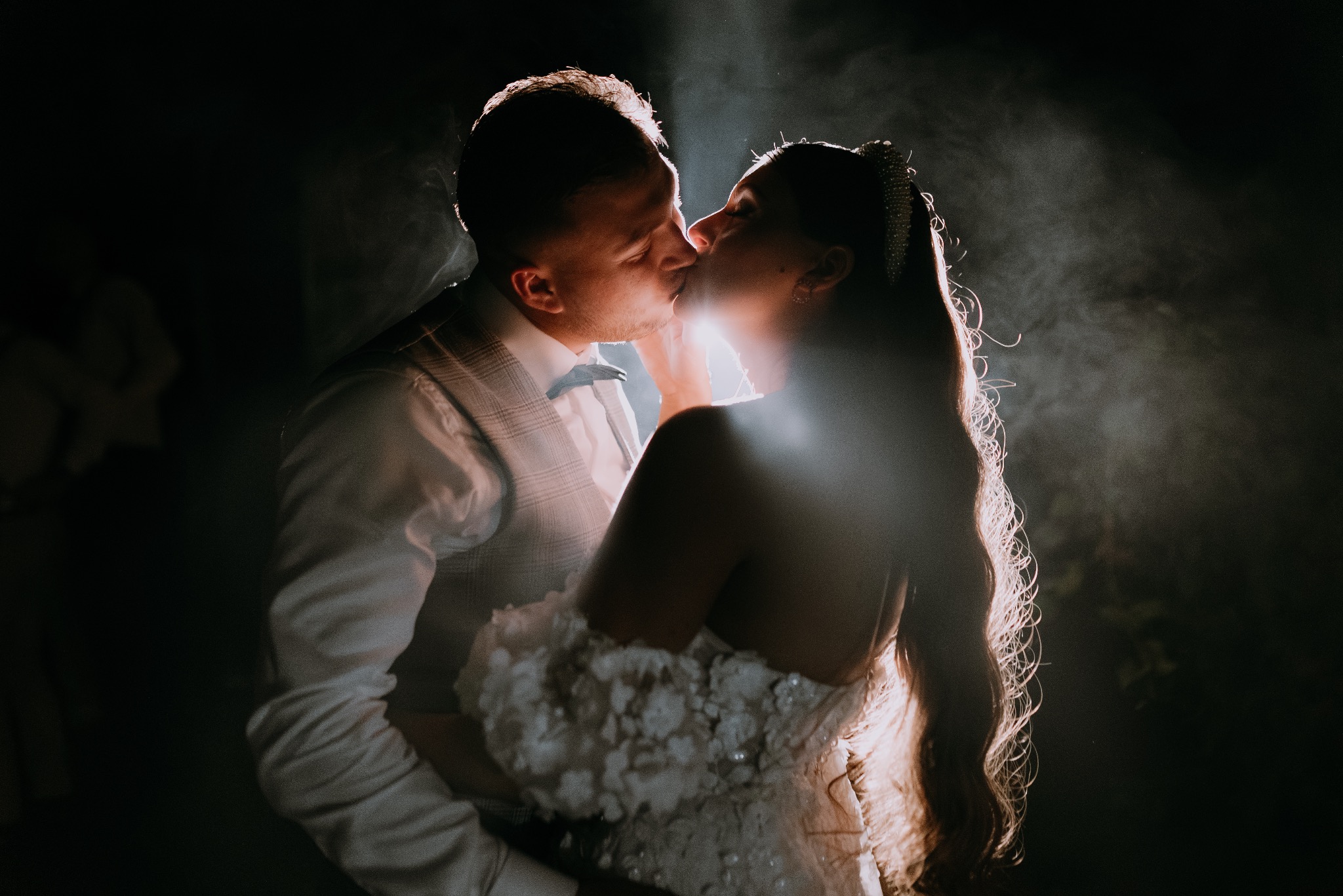 Para młoda w pocałunku, oświetlona od tyłu, on w kamizelce w kratę i muszce, ona w sukni z aplikacjami kwiatowymi i diademem we włosach, ciemne tło.