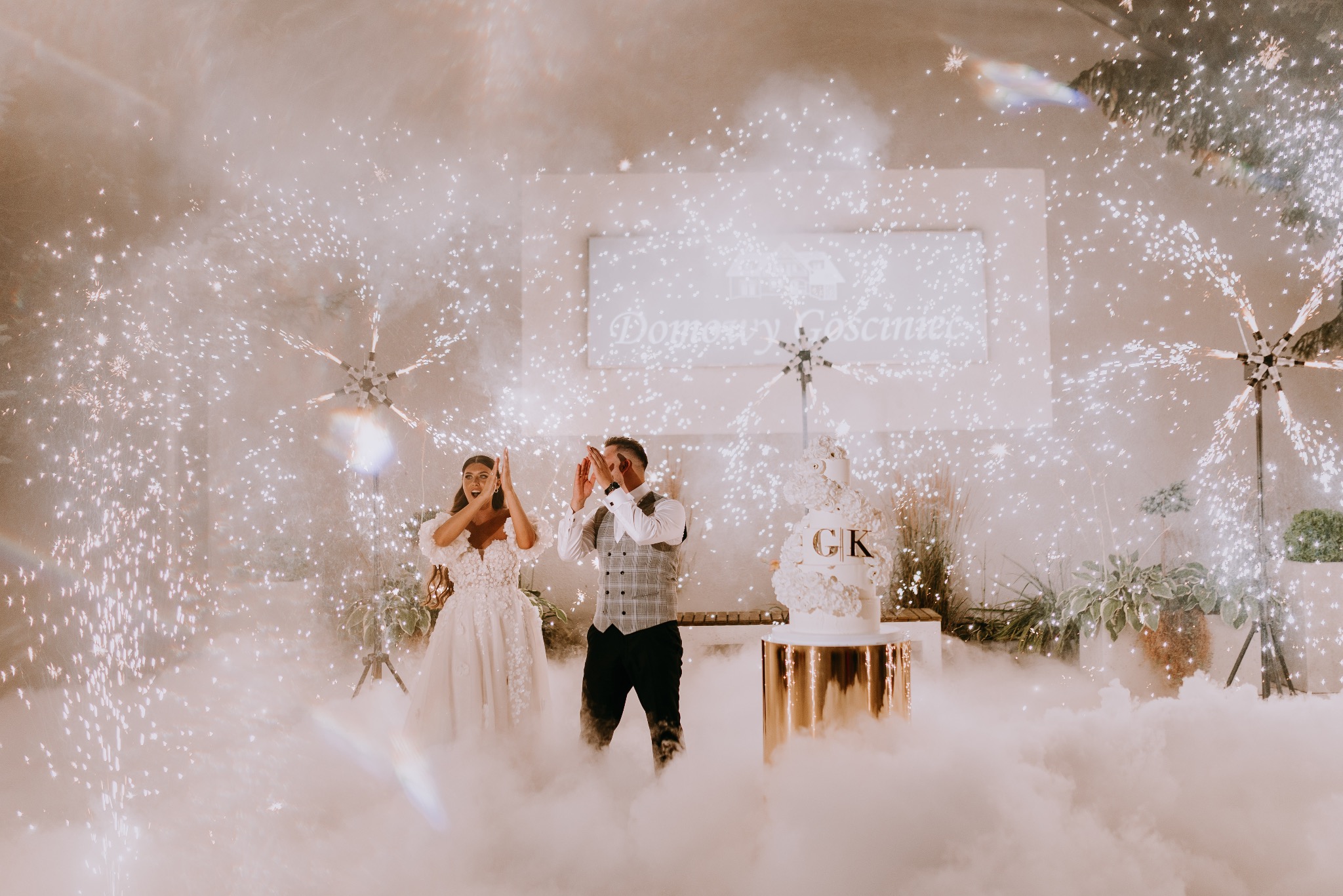 Para młoda, kobieta w białej sukni z kwiatami i mężczyzna w szarej kamizelce, klaszczą w dłonie podczas pokazu fajerwerków w pomieszczeniu, obok tort weselny z inicjałami 'GK', dym i światła tworzą...