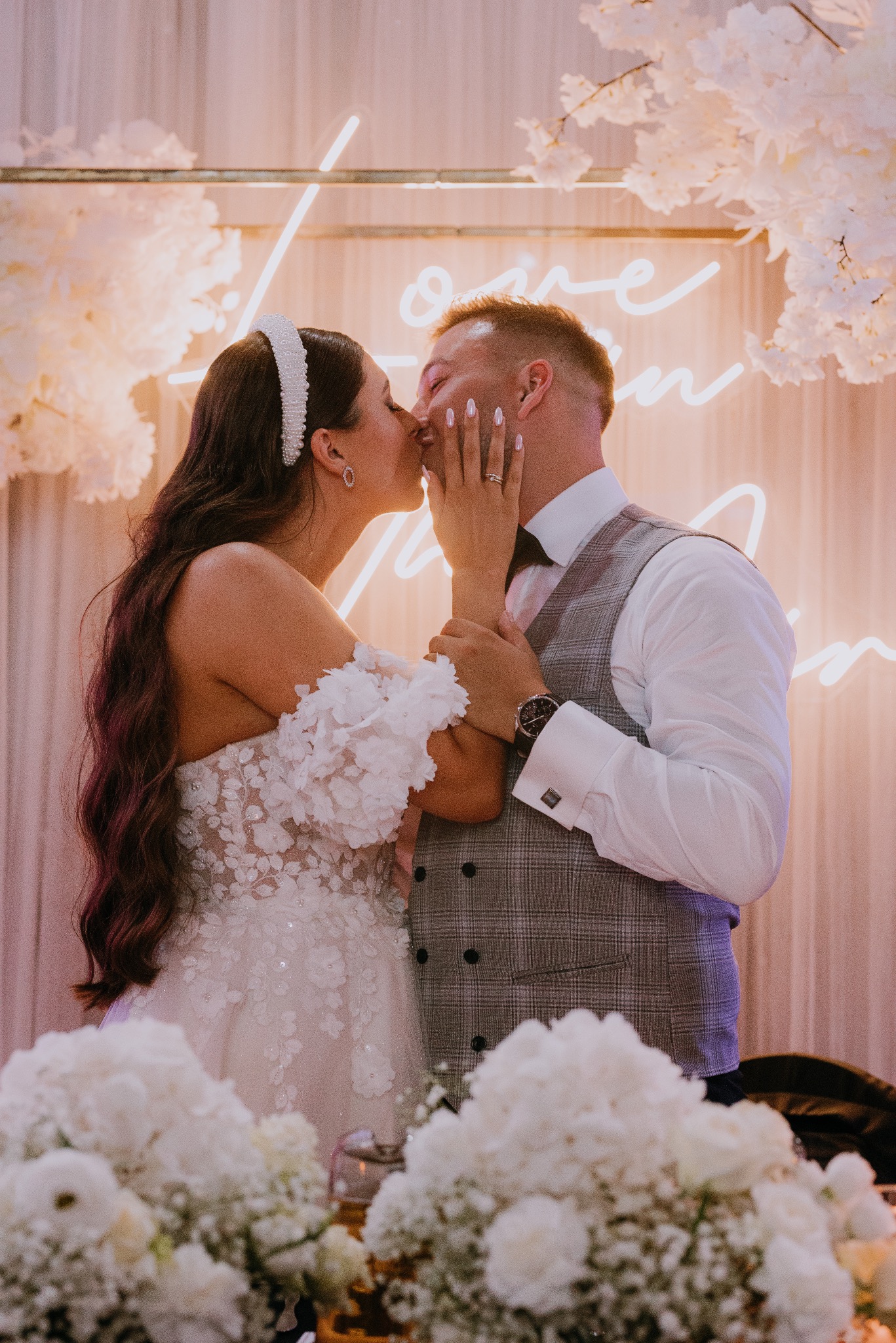 Pocałunek nowożeńców na tle neonowego napisu 'Love Begins', bukiety białych kwiatów w pierwszym planie, panna młoda w sukni z kwiatowymi aplikacjami i perłową opaską, pan młody w kraciastej...