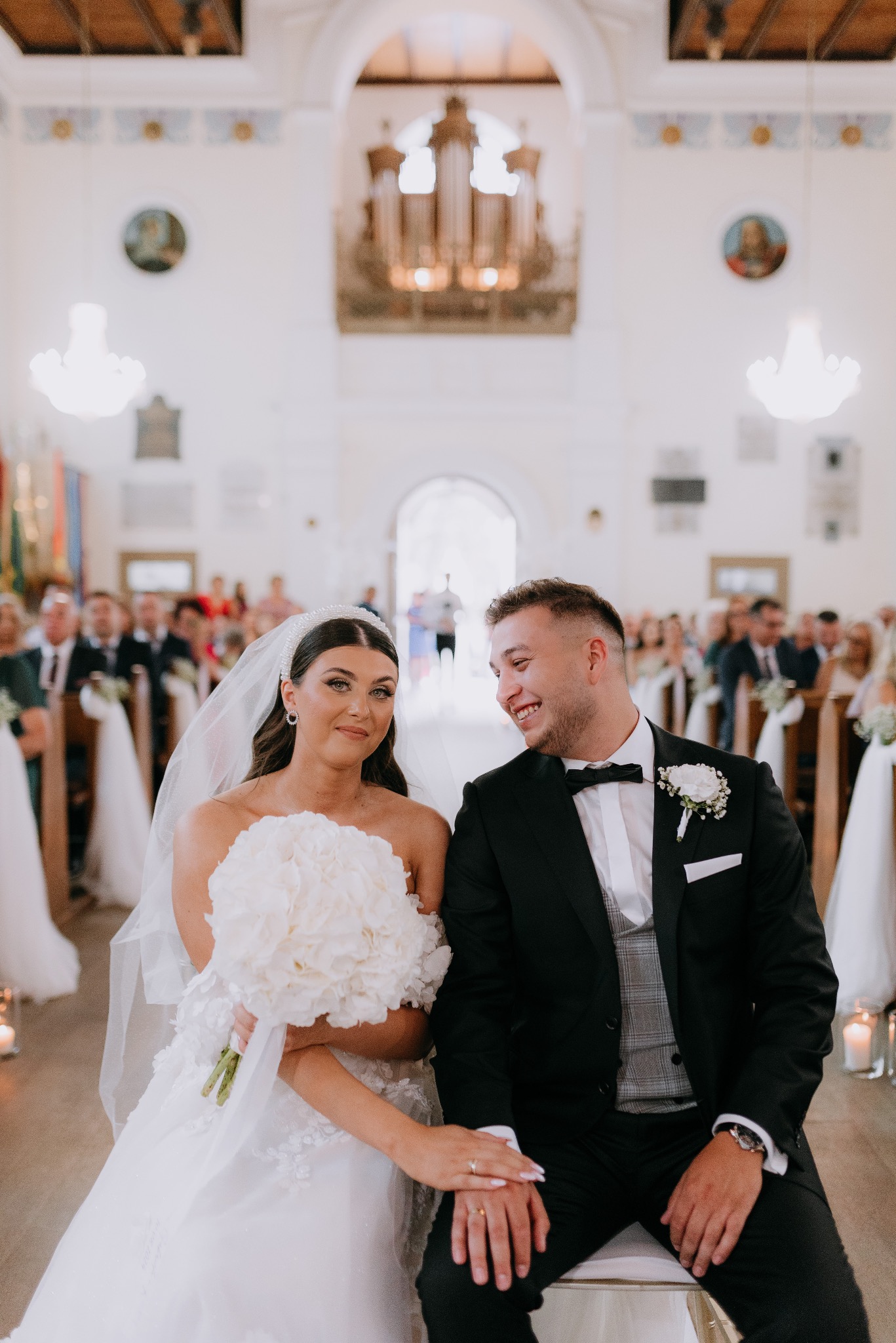 Panna młoda w koronkowej sukni z bukietem białych kwiatów i pan młody w czarnym smokingu siedzą w kościele podczas ceremonii ślubnej, w tle organy i goście.