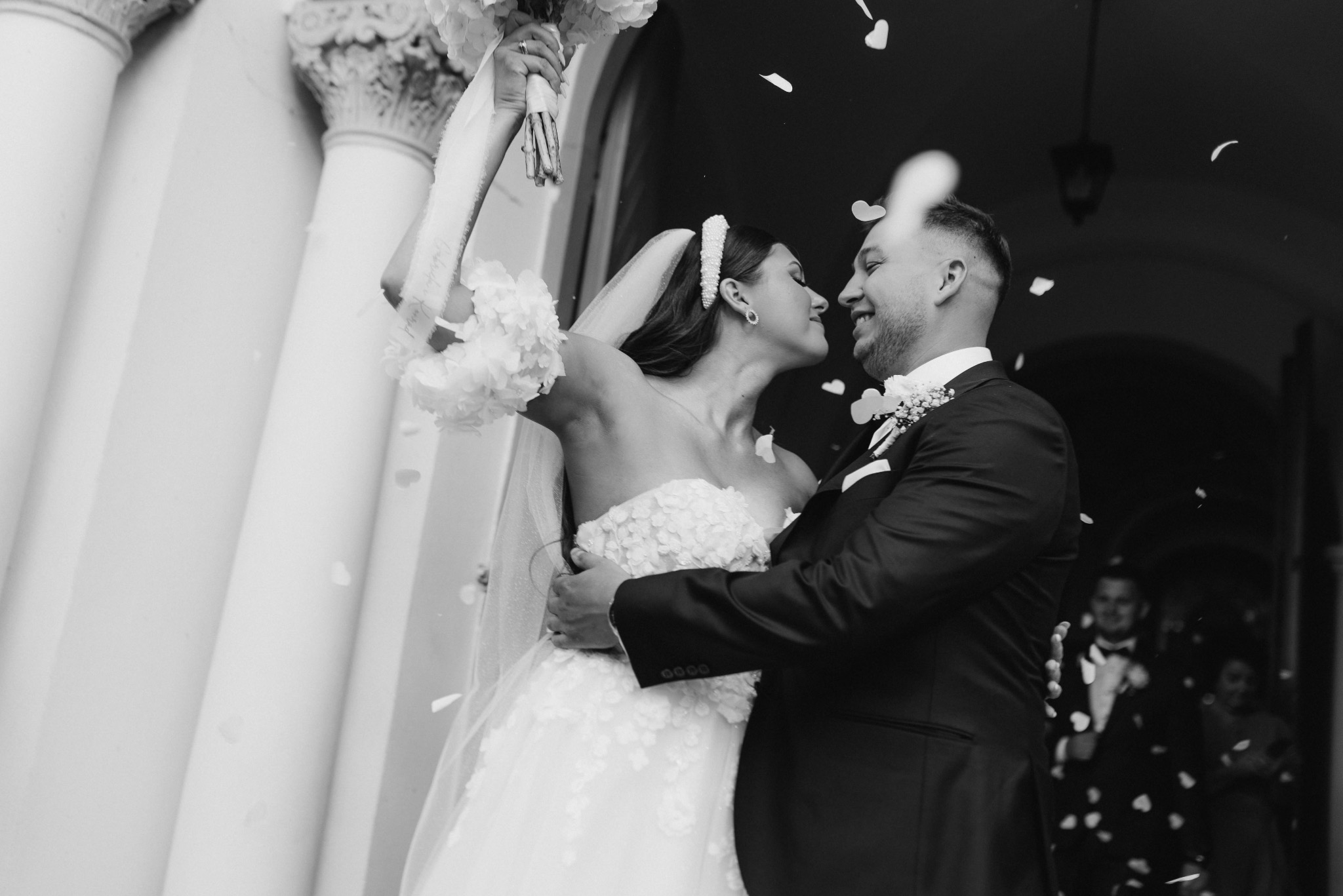 Czarno-białe zdjęcie nowożeńców w objęciach, obsypywanych płatkami, z kolumnami w tle i gośćmi weselnymi w oddali.