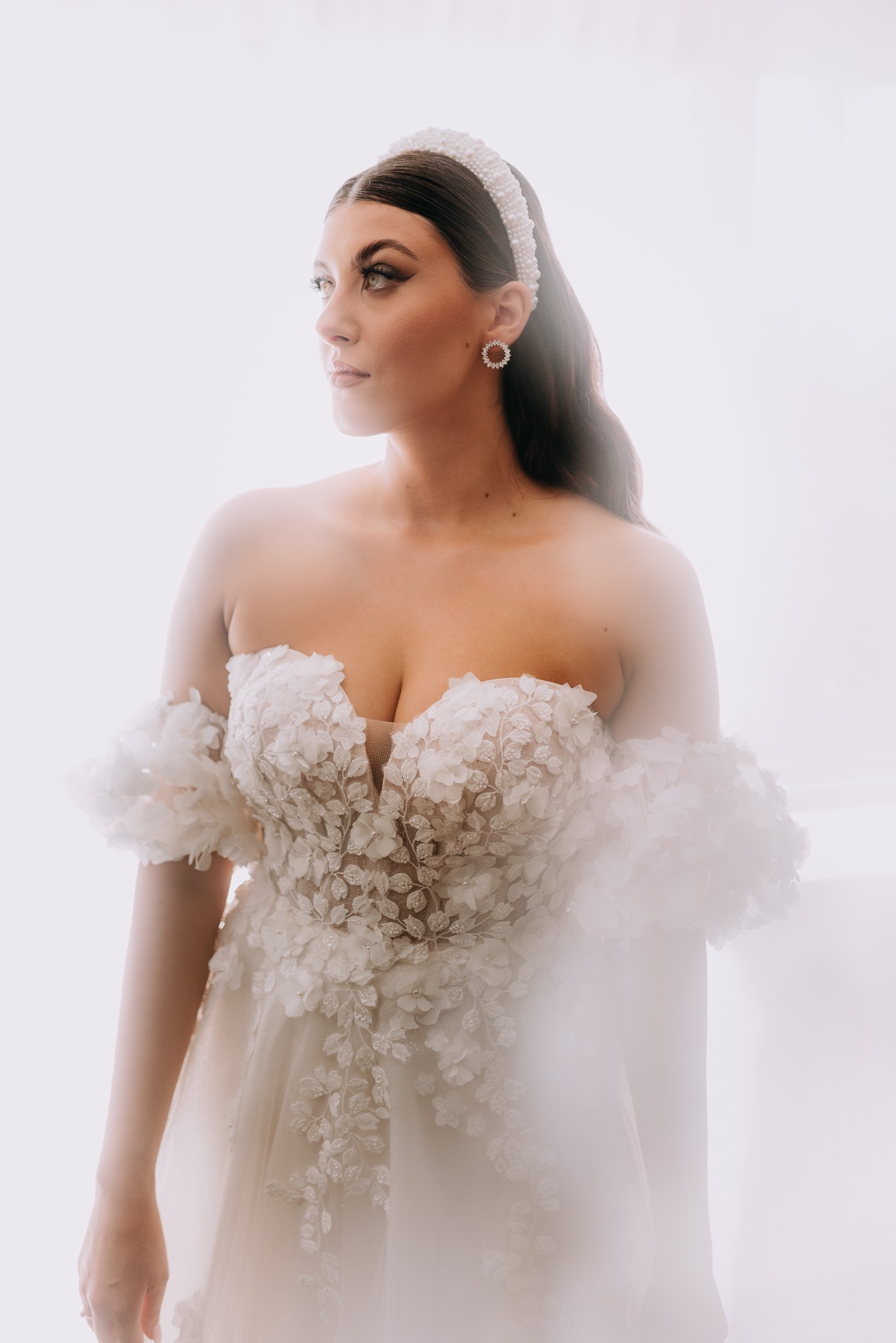 Panna młoda w sukni ślubnej z kwiatowymi aplikacjami, perłową opaską i kolczykami w kształcie rozet, patrzy w bok na jasnym tle.