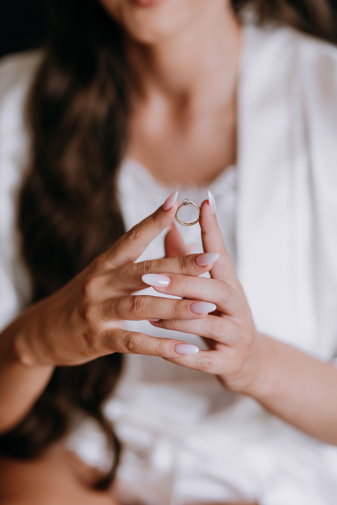 Dłonie kobiety z manicure trzymają złoty pierścionek zaręczynowy z brylantem na tle białego szlafroka.