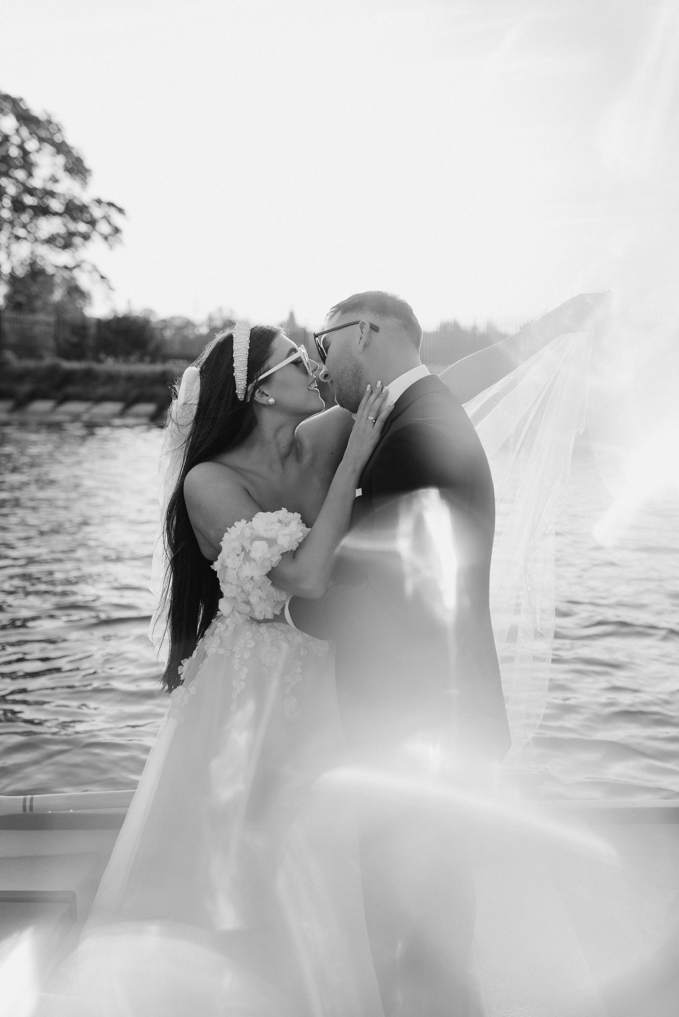 Czarno-białe zdjęcie całującej się pary młodej na łodzi; panna młoda w sukni z aplikacjami kwiatowymi i okularach w stylu retro, pan młody w garniturze i okularach, w tle woda i zieleń.