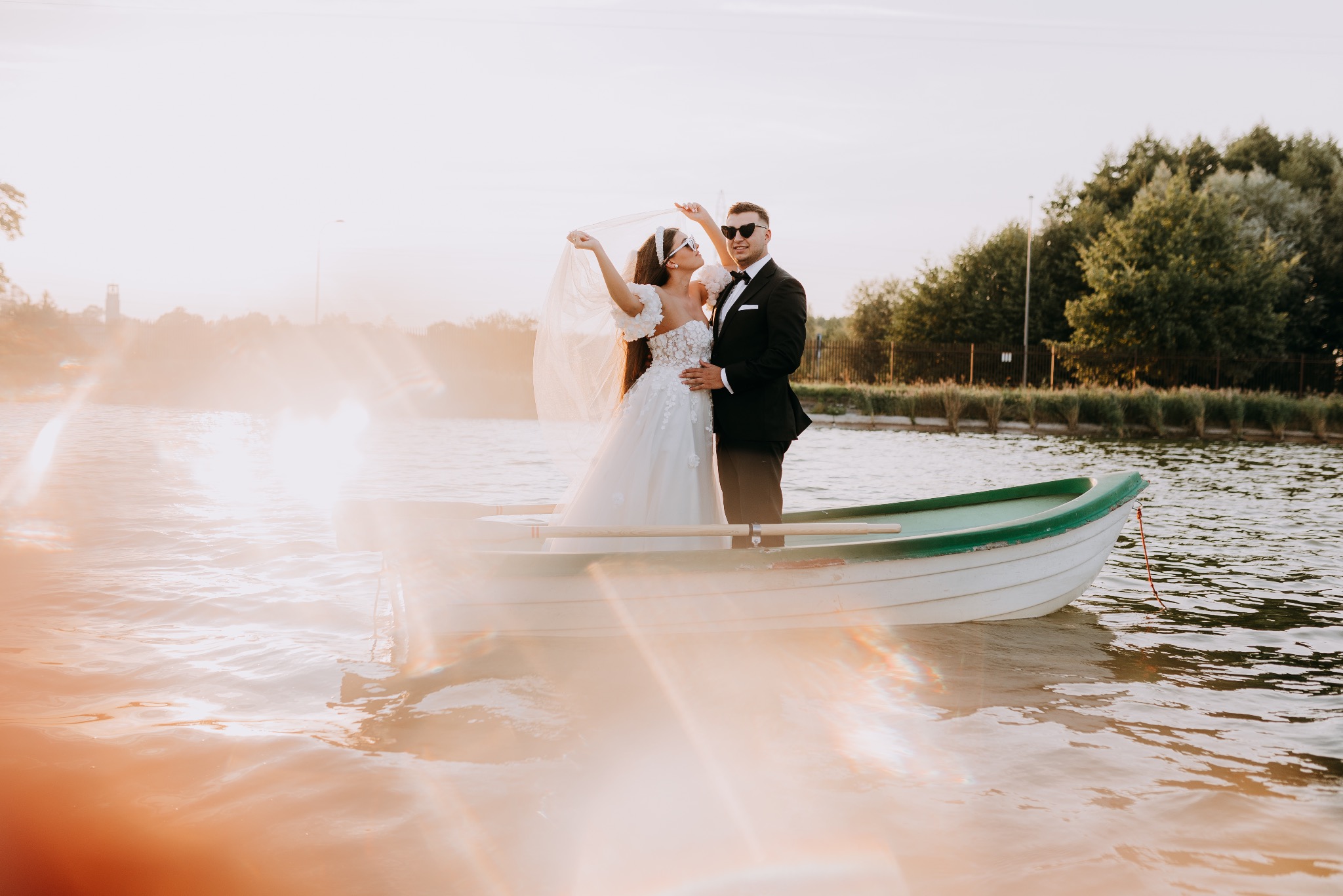 Para młoda w łódce na jeziorze w promieniach zachodzącego słońca. Panna młoda w białej sukni z kwiatowymi aplikacjami i welonem, pan młody w czarnym smokingu i okularach przeciwsłonecznych.