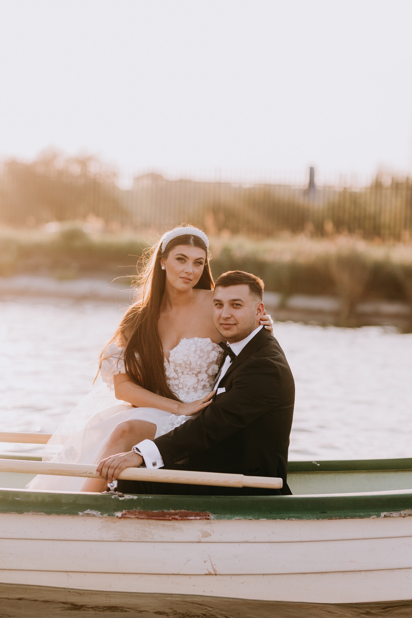 Para młoda w łódce na jeziorze podczas zachodu słońca, panna młoda w sukni z kwiatowym wzorem i diademem, pan młody w czarnym smokingu.
