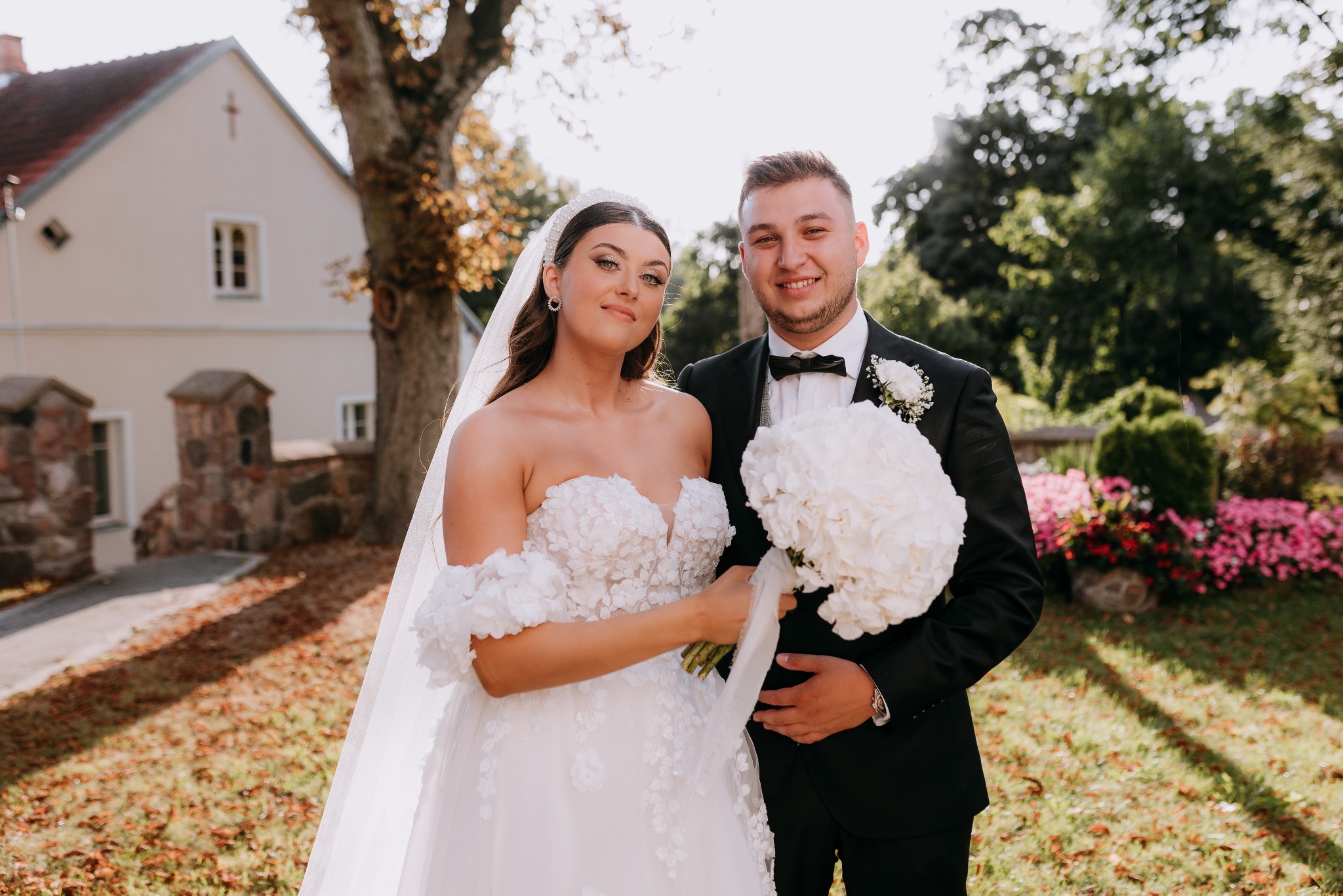 Para młoda w plenerze: panna młoda w sukni z kwiatowymi aplikacjami i welonem, pan młody w czarnym smokingu z bukietem białych kwiatów, w tle budynek i zieleń.