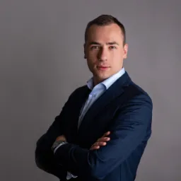 Portret mężczyzny w granatowym garniturze i błękitnej koszuli, z zegarkiem na lewym nadgarstku, pozującego na szarym tle z założonymi rękami.