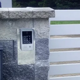 Nowoczesny wideodomofon z klawiaturą numeryczną zamontowany na kamiennym słupku ogrodzenia z białą, poziomą bramą w tle.