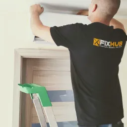 Instalator w czarnej koszulce z logo firmy FIXHUB, montuje białą jednostkę klimatyzacyjną na ścianie, stojąc na aluminiowej drabinie z zielonymi elementami, widoczny fragment drzwi.