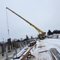 F.H.U. Monika Chojnacka - Żółty dźwig samochodowy na zasnieżonym placu budowy podnosi szalunek w pobliżu zbrojonych fundamentów budynku żelbetowego, widoczne elementy konstrukcyjne i zbrojenia.