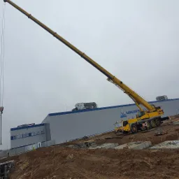 Żółty dźwig teleskopowy w Rawa Mazowiecka podnosi element konstrukcyjny na placu budowy, w tle szary budynek z napisem, pochmurne niebo.