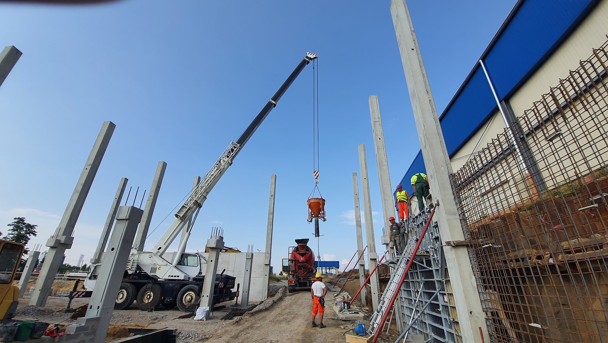 Budowa hali: żelbetowe słupy, dźwig podnosi pojemnik z betonem, zbrojenie ścian w tle, pracownicy w kaskach i odblaskowych ubraniach na rusztowaniu.