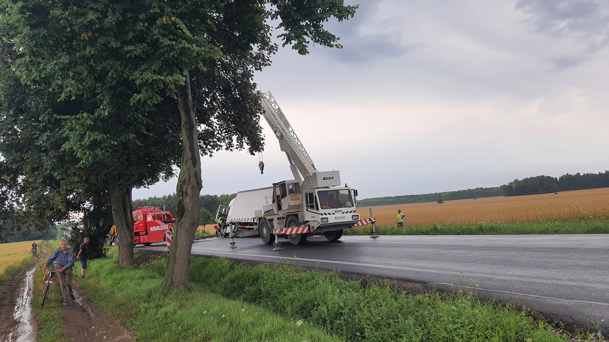 Biały samochód ciężarowy z wysięgnikiem na poboczu asfaltowej drogi, obok pola zboża i drzew, w tle czerwony samochód ciężarowy, pochmurne niebo.