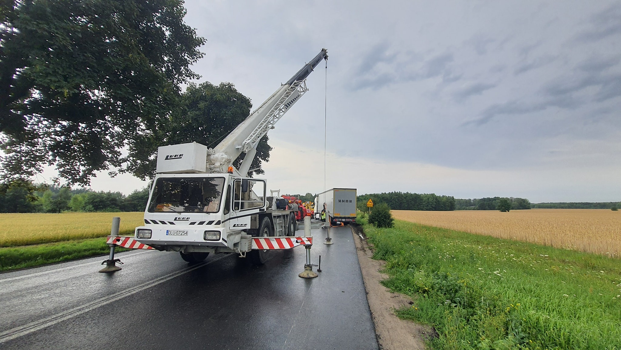 Biały dźwig samochodowy marki Liebherr z wysuniętym ramieniem na mokrej, asfaltowej drodze, w tle pole zboża i zaparkowana ciężarówka z naczepą.
