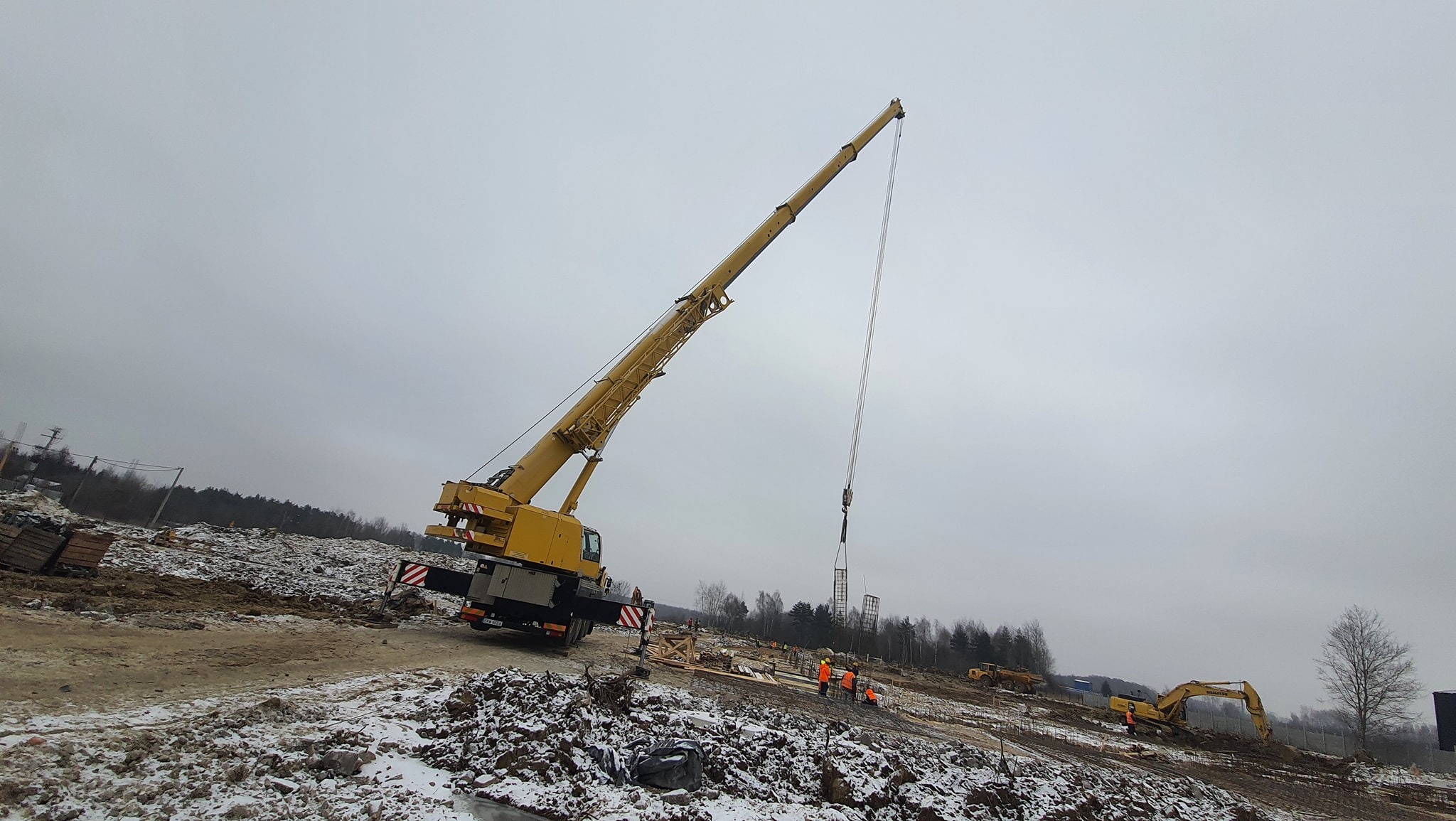 Żółty dźwig samochodowy podnosi metalowy szkielet zbrojenia na placu budowy w pochmurny dzień, w tle widoczne inne maszyny budowlane i pracownicy w pomarańczowych kamizelkach.