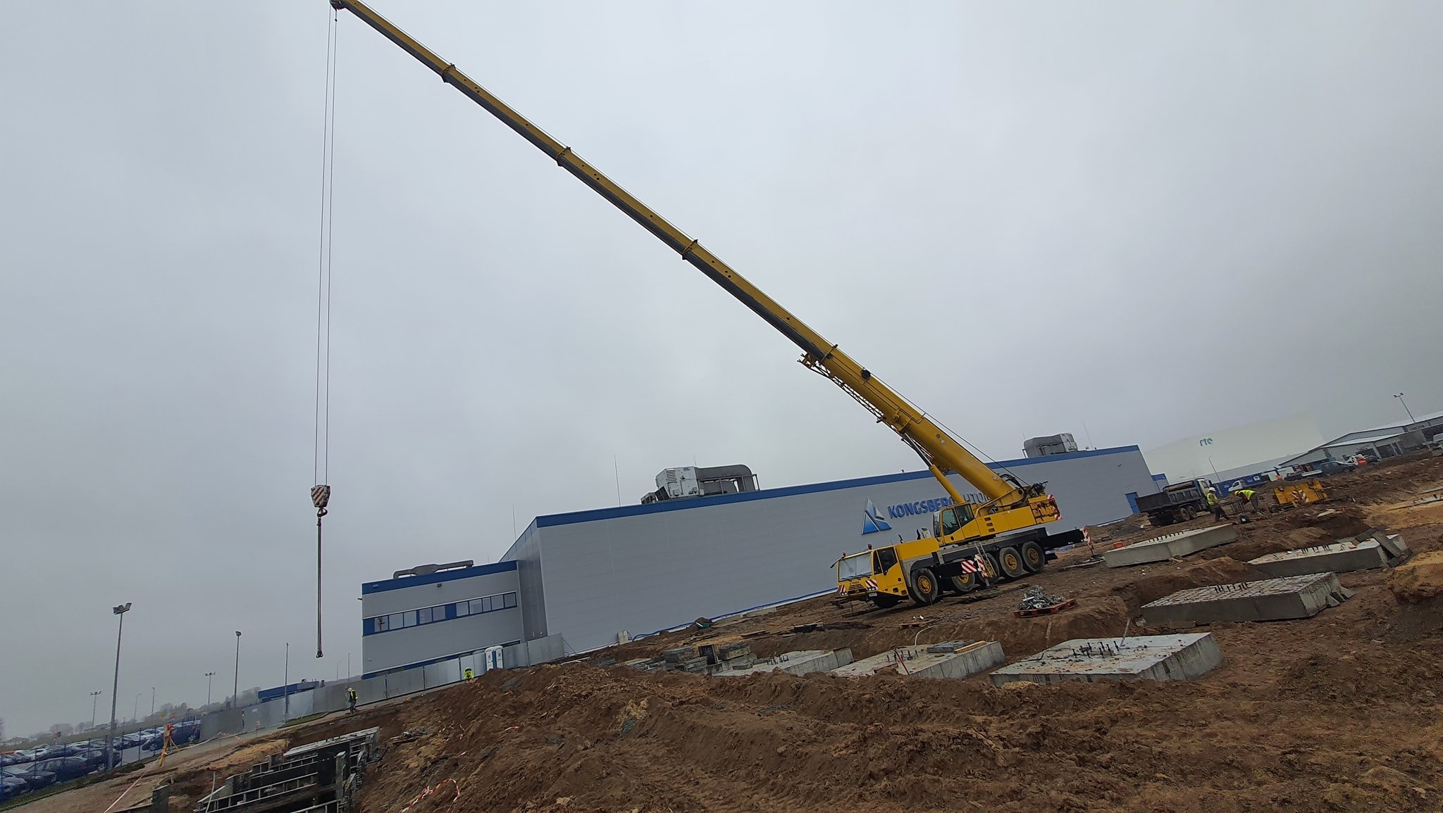 Żółty dźwig teleskopowy w Rawa Mazowiecka podnosi element konstrukcyjny na placu budowy, w tle szary budynek z napisem, pochmurne niebo.