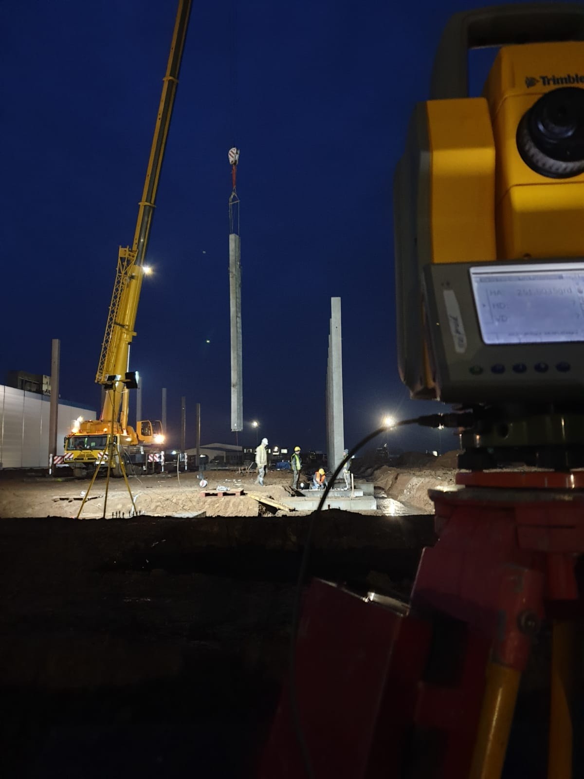 Montaż prefabrykowanych słupów żelbetowych na placu budowy w nocy, widoczny żółty dźwig i pracownicy w kaskach, w pierwszym planie dalmierz geodezyjny Trimble.