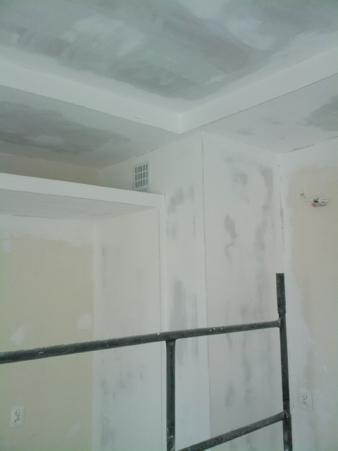 Wnętrze w trakcie wykończenia: sufit z widocznymi łączeniami płyt gipsowo-kartonowych przed szpachlowaniem, fragment rusztowania, ściany z widocznymi śladami po szpachlowaniu, kratka wentylacyjna...