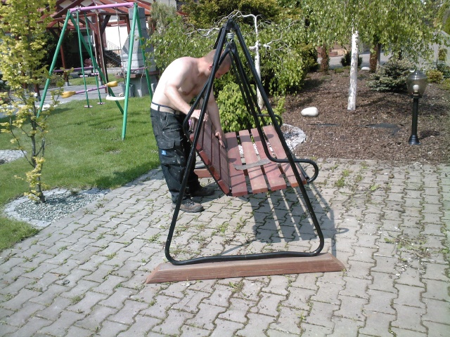 Mężczyzna bez koszuli montuje drewnianą huśtawkę ogrodową na tle zadbanego trawnika i drzew, widoczna kostka brukowa i plac zabaw w oddali.