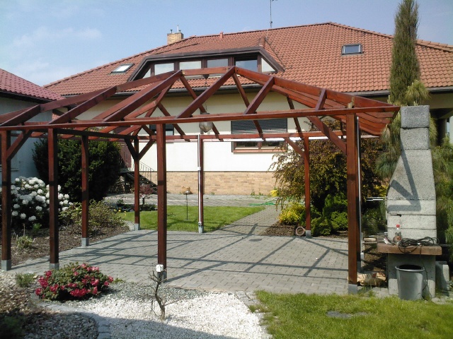 Drewniana konstrukcja altany z widoczną więźbą dachową, na tle domu z czerwoną dachówką i murowanego grilla z akcesoriami, otoczona zielenią ogrodu i brukowanym tarasem.