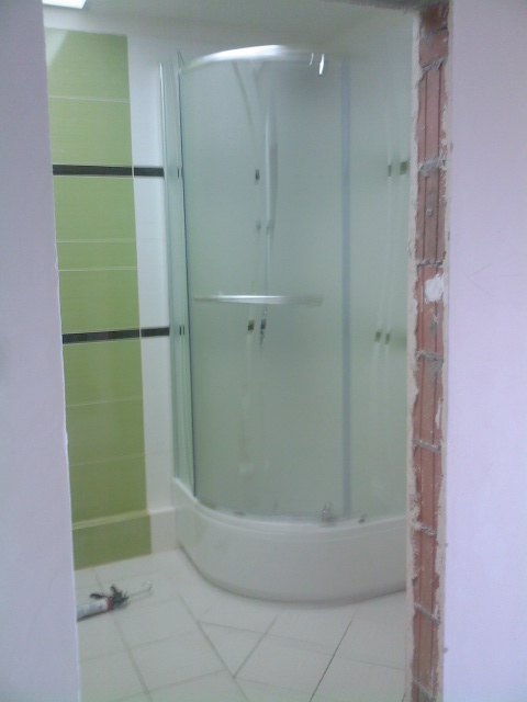 Narożna kabina prysznicowa z półprzezroczystymi szybami, widoczna w trakcie montażu, z narzędziem do silikonowania na podłodze i surowym wykończeniem otworu drzwiowego.