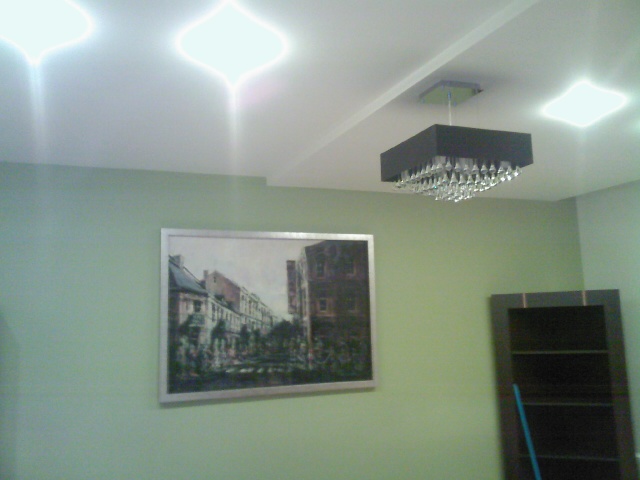 Czarny, kwadratowy żyrandol z kryształkami zawieszony na białym suficie z trzema wbudowanymi lampami, ściana pomalowana na jasnozielono z obrazem w srebrnej ramie przedstawiającym uliczkę miejską...