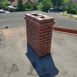 Nowy, ceglany komin na płaskim dachu z papy, widok z góry, w tle zabudowa jednorodzinna.