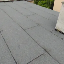 Szary dach pokryty papą termozgrzewalną z widocznym kominem i fragmentami budynków w tle.