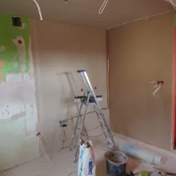 Wnętrze pokoju w trakcie prac wykończeniowych: ściany pokryte świeżym tynkiem, widoczne instalacje elektryczne i hydrauliczne, drabina, worek z zaprawą i narzędzia murarskie na podłodze.