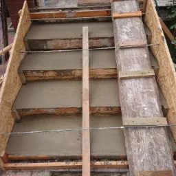 Budowa schodów zewnętrznych: szalunek z desek wypełniony świeżym betonem, wzmocniony stalowymi prętami, widoczne fragmenty cegieł i podłoża.