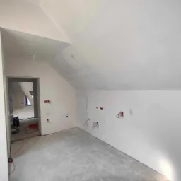 Poddasze w trakcie prac wykończeniowych, świeżo pomalowane ściany na biało, widoczne wystające kable elektryczne, surowa podłoga i otwór drzwiowy do kolejnego pomieszczenia.
