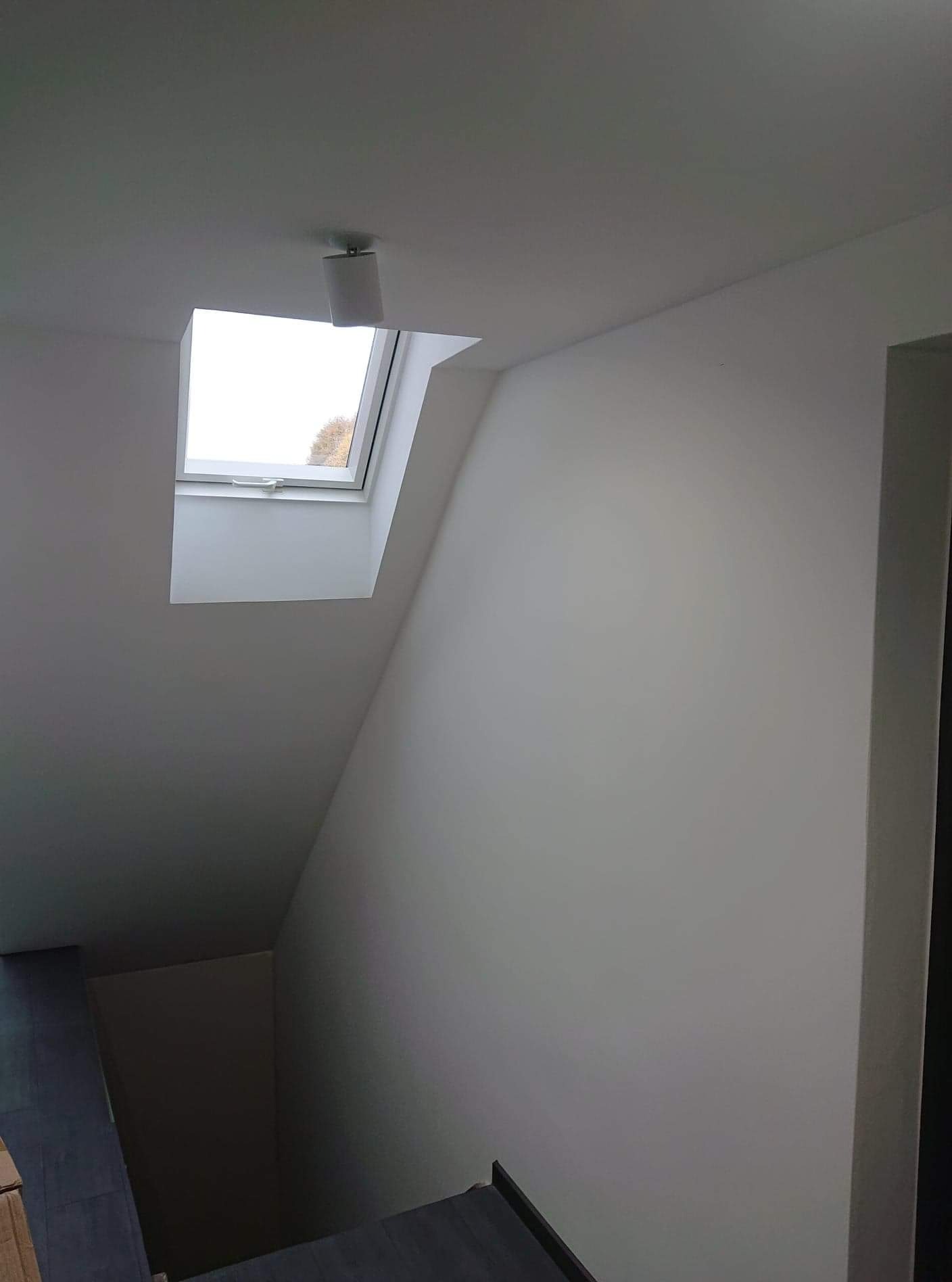 Poddasze po malowaniu na biało, widoczne okno dachowe z zamontowaną lampą punktową i fragment schodów z szarymi stopniami.