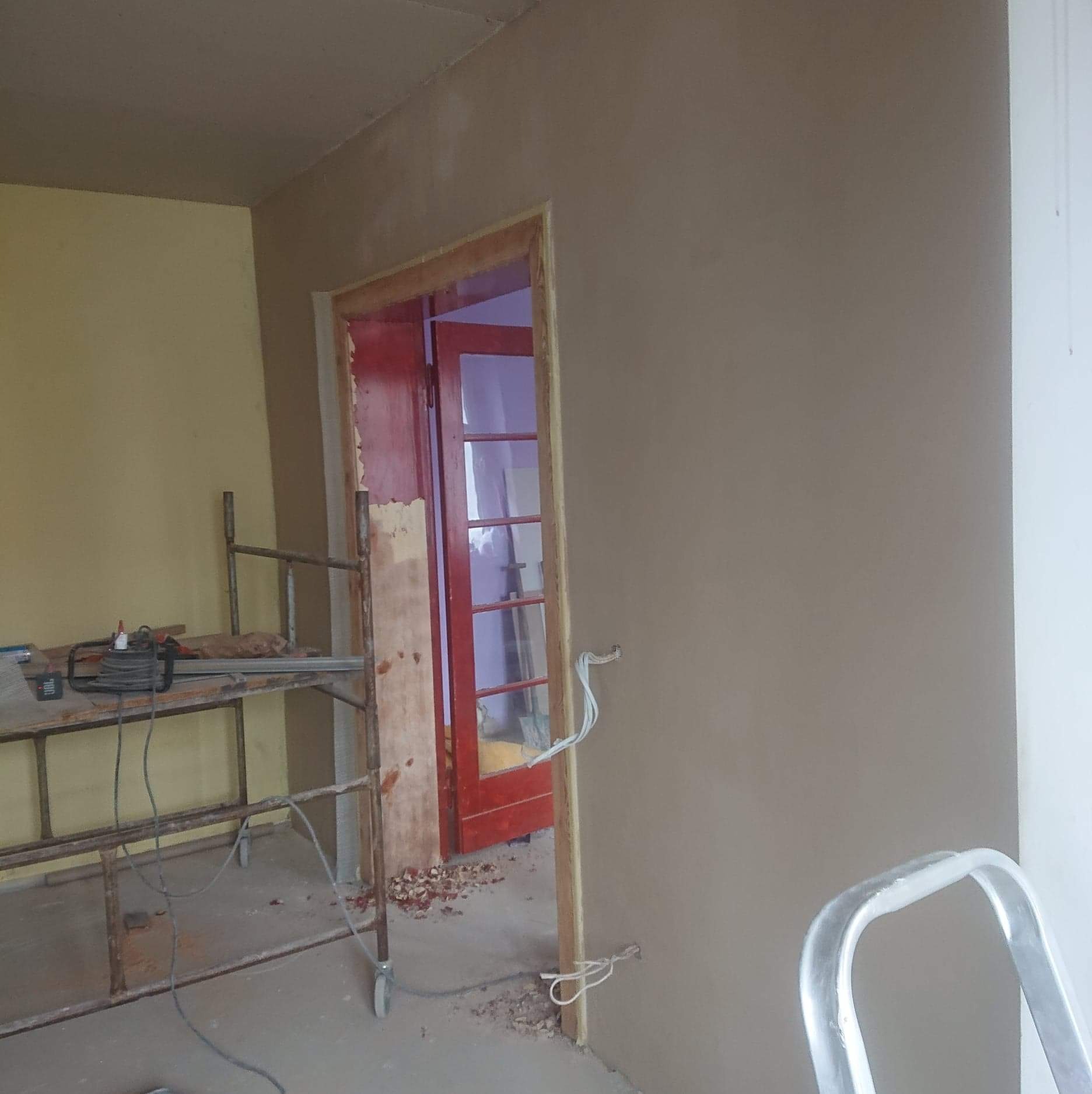 Wnętrze w trakcie prac wykończeniowych: ściana pokryta świeżym tynkiem w odcieniach beżu, widoczne kable wystające ze ściany, fragment rusztowania z narzędziami, czerwone drzwi z szybami w tle...