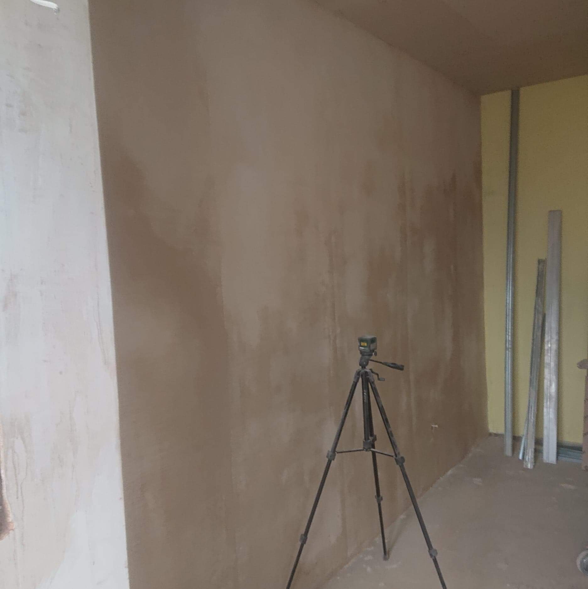 Świeżo otynkowana ściana z widocznym laserowym niwelatorem na statywie, profile aluminiowe opierają się o ścianę w tle.