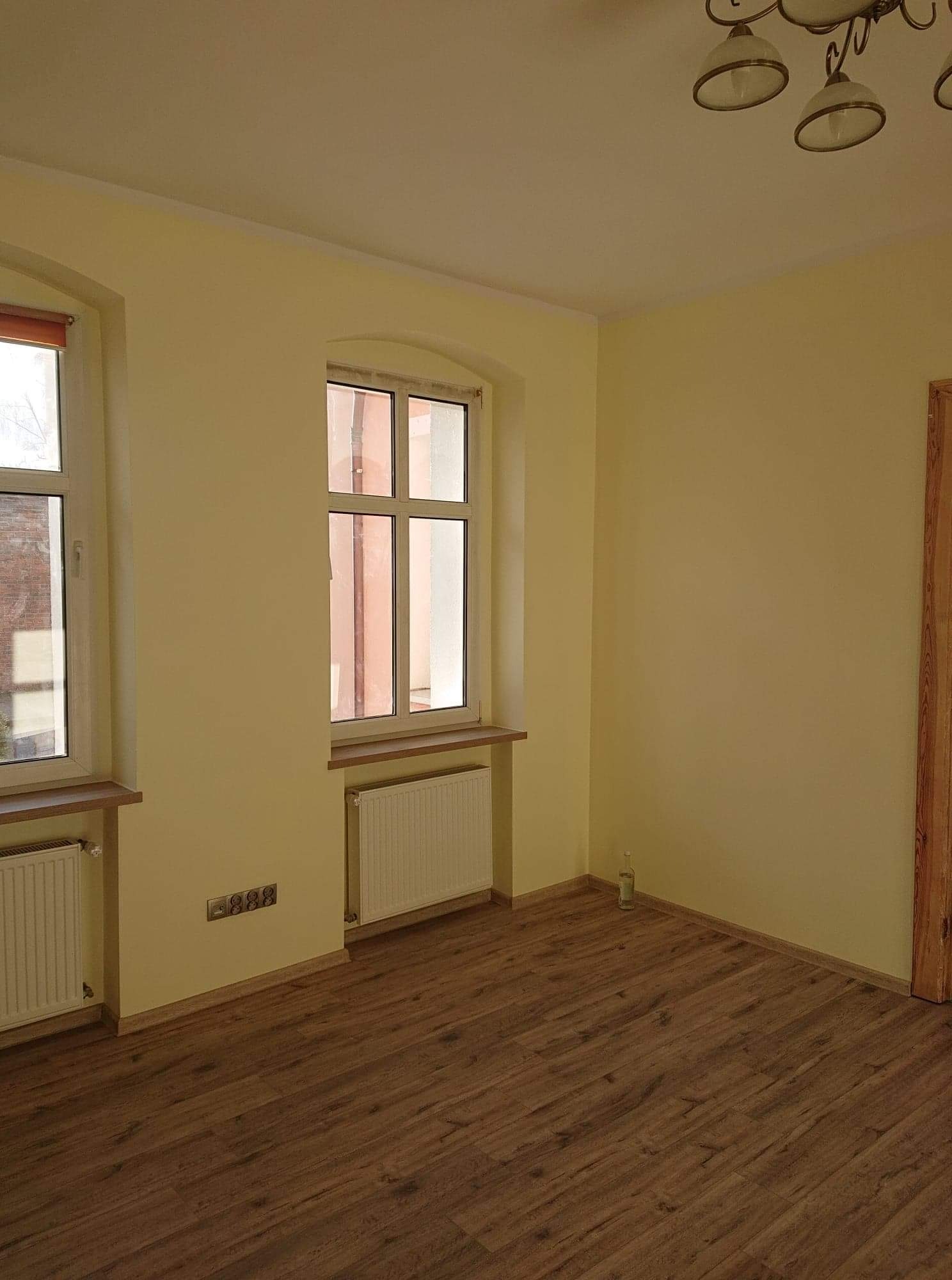 Pomalowane na jasnożółto puste pomieszczenie z dwoma oknami, drewnianą podłogą i białymi grzejnikami.