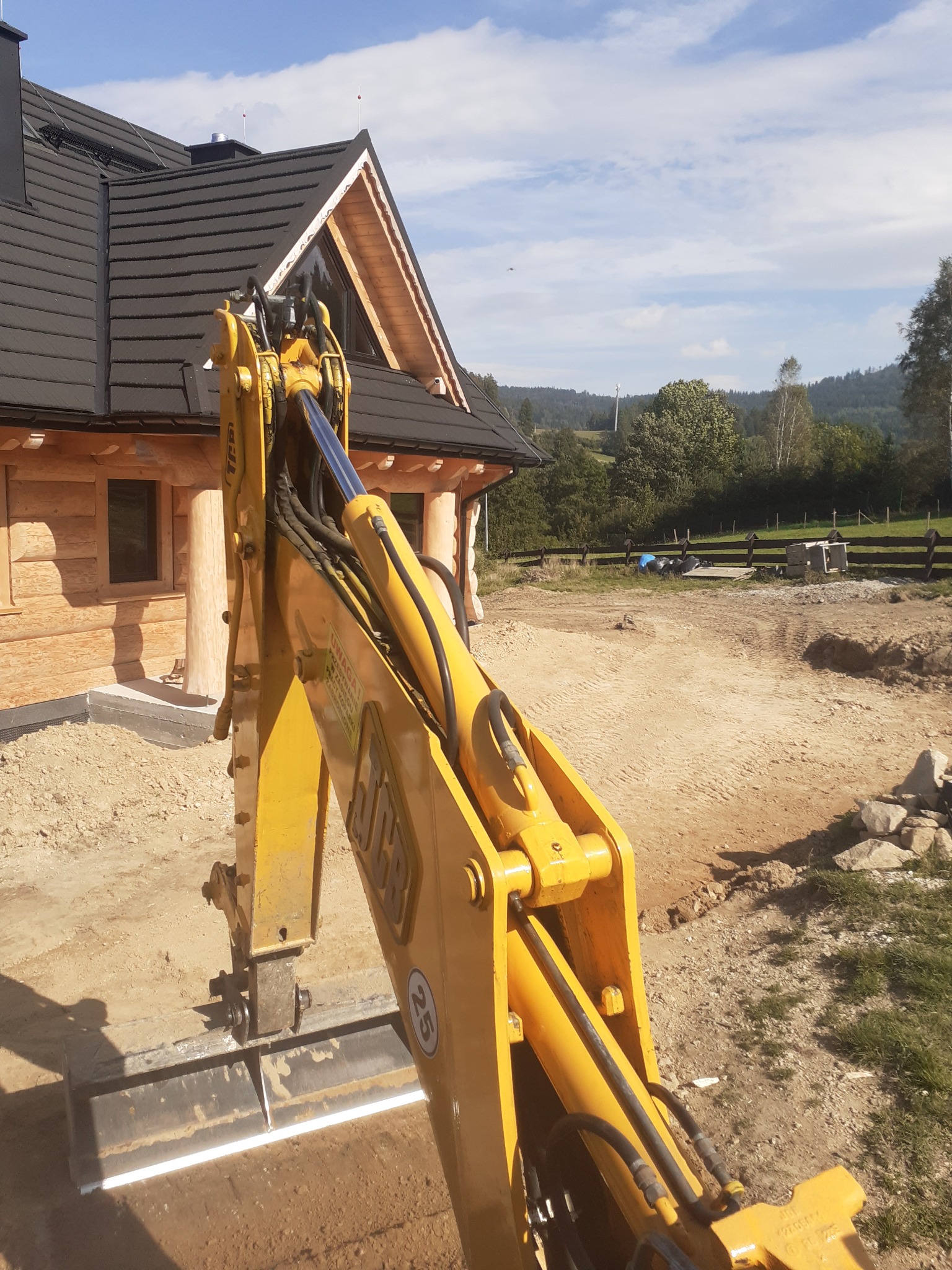 Żółta koparko-ładowarka JCB z łyżką równającą na placu budowy przed drewnianym domem z ciemnym dachem, w tle pagórkowaty krajobraz.