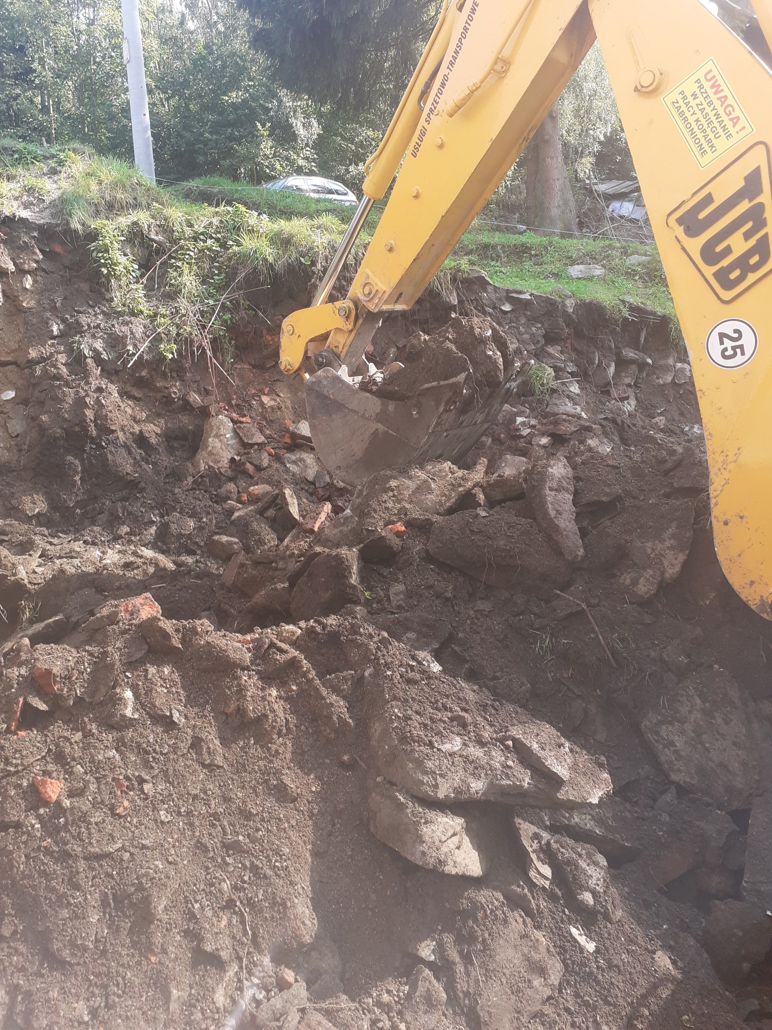 Żółta koparka JCB w trakcie pracy, łyżka zagłębia się w ziemię z gruzem i fragmentami cegieł, widoczny numer 25 na maszynie, w tle zieleń i drzewa.