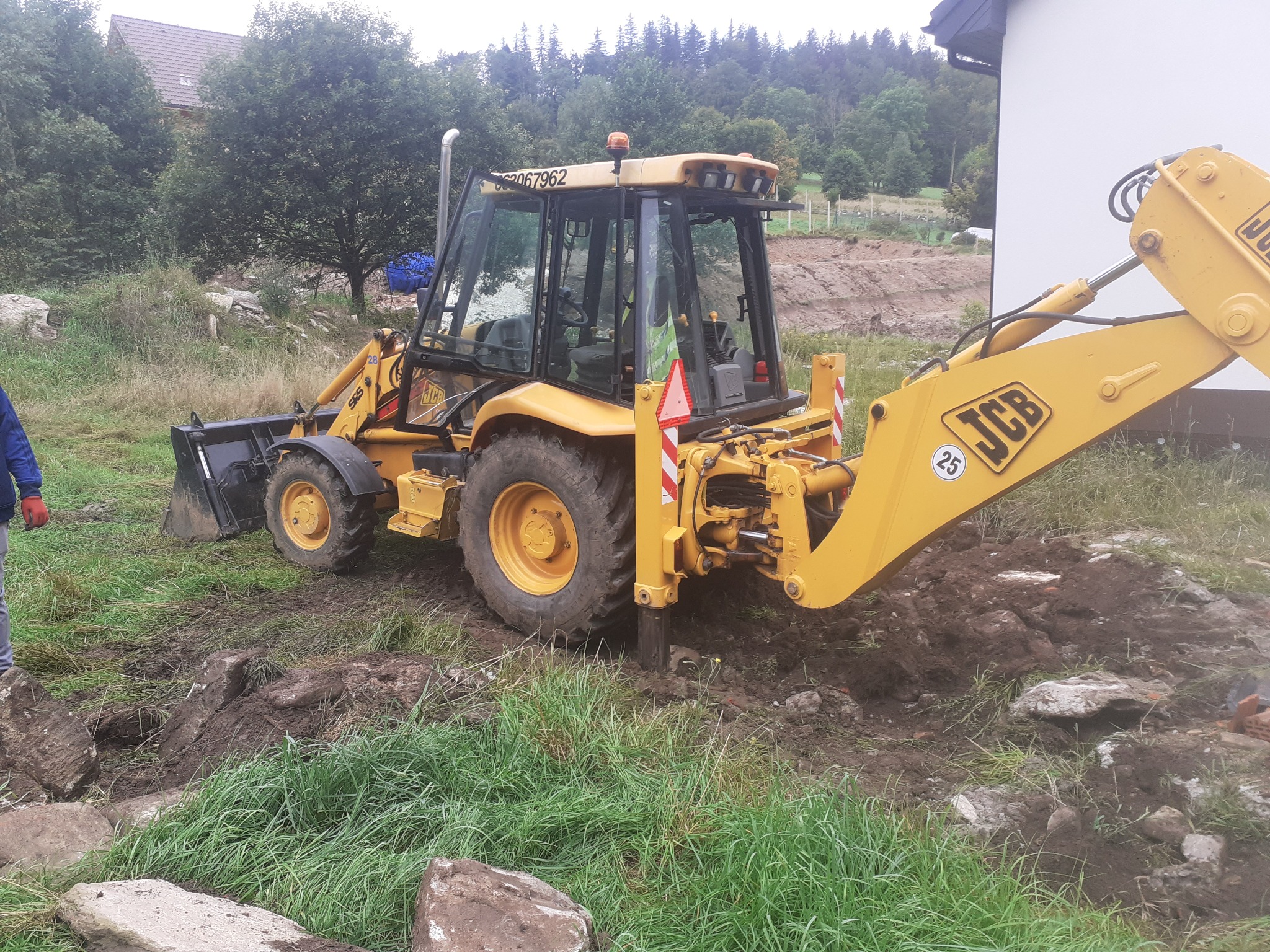 Żółta koparko-ładowarka JCB w trakcie prac ziemnych na terenie w Stronie Śląskie, widoczny wykop z kamieniami i fragment budynku w tle.