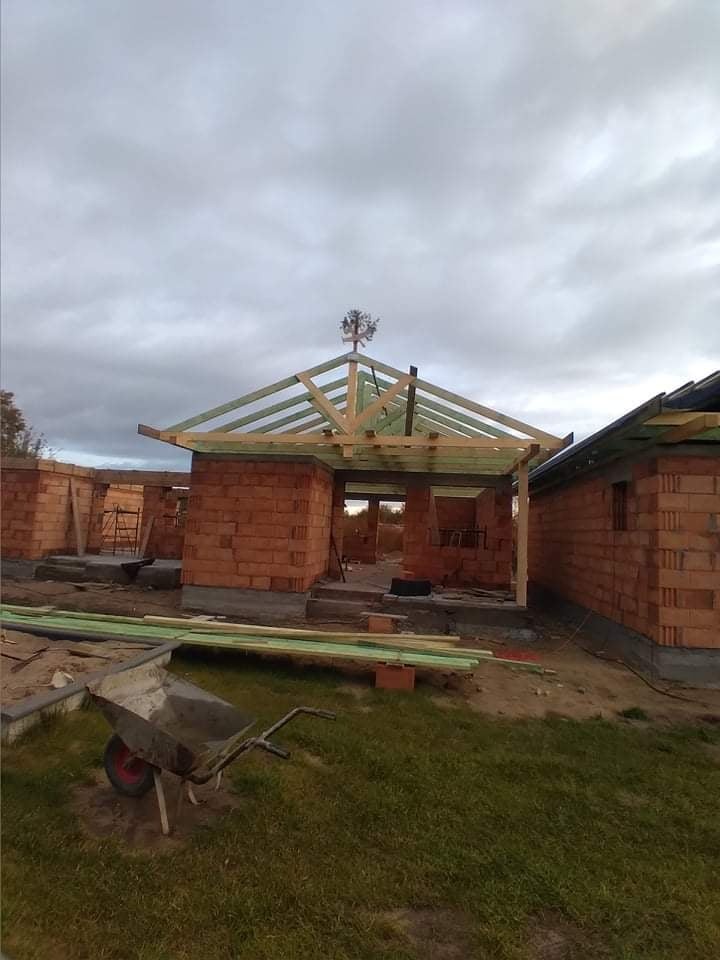 Budowa domu z czerwonej cegły, widoczna konstrukcja dachu z drewnianych belek, w tle pochmurne niebo, na szczycie dachu dekoracyjny element metalowy, na pierwszym planie taczka i deski.