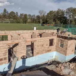 Wznoszenie ścian parterowego domu z czerwonej cegły, widoczne zbrojenia i fundamenty, w tle zielone boisko sportowe i drzewa.