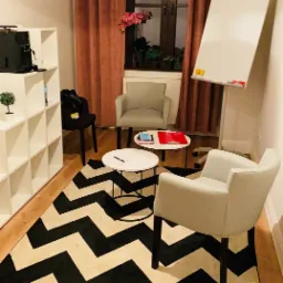 Jasne wnętrze gabinetu z dwoma fotelami, stolikami kawowymi, flipchartem i wzorzystym dywanem w zygzaki.