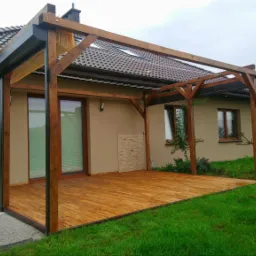 Zadaszona drewniana konstrukcja tarasowa przylegająca do beżowego domu jednorodzinnego z drewnianą podłogą, widok z zewnątrz na ogród z trawą i sąsiedni dom z panelami słonecznymi na dachu.