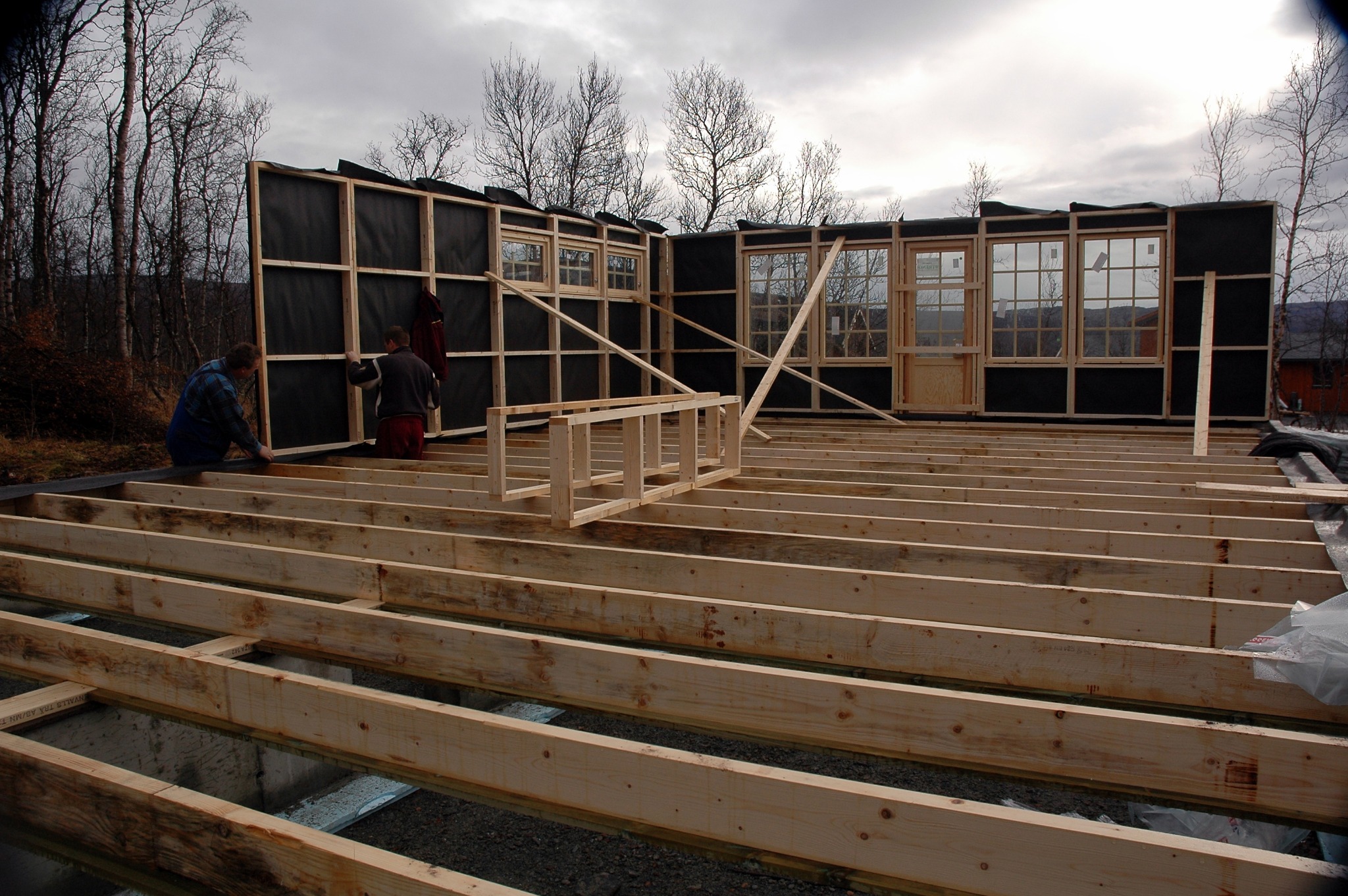 Budowa domu w trakcie: widoczny szkielet drewniany z oknami i drzwiami, izolacja, fundament z belek, dwóch robotników przy pracy, pochmurne niebo w tle.